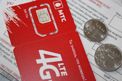 A close-up of a red SIM card holder from MTS with its accompanying packaging. The card displays '4G LTE' and includes a microchip design. Two coins are placed on top of a printed document, partially obscuring the text in Russian.