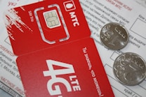 A close-up of a red SIM card holder from MTS with its accompanying packaging. The card displays '4G LTE' and includes a microchip design. Two coins are placed on top of a printed document, partially obscuring the text in Russian.