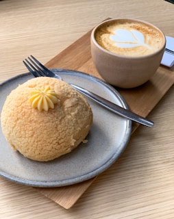 A light brown, crumbly pastry with a small decorative dollop of cream on top is placed on a gray plate. Next to it is a fork. A latte with a heart-shaped milk froth design is served in a beige cup on a wooden board.