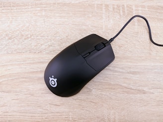 A black computer mouse with a cord rests on a light wooden surface. The mouse features a minimalist design and a logo near its back.
