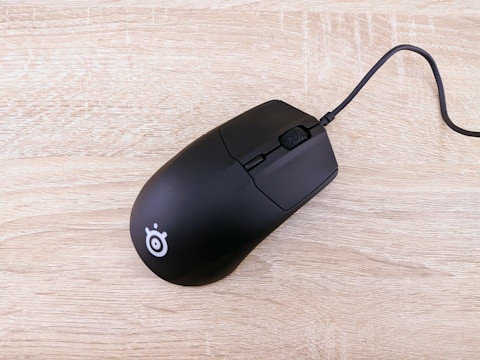 A black computer mouse with a cord rests on a light wooden surface. The mouse features a minimalist design and a logo near its back.