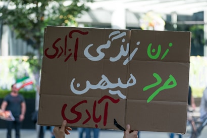 A person holds a cardboard sign with text written in Persian script. The sign features large letters in colors such as green, white, and red. The background includes blurred figures and trees, indicating an outdoor setting.