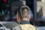 A person with short hair is wearing a reflective safety vest. The background includes blurred colors and shapes, possibly flags, and an urban setting with cars.