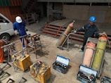 Two people wearing protective gear, including helmets, are working with welding equipment. They are surrounded by various tools and large gas cylinders on carts, likely for industrial use. The setting appears to be an industrial workshop with metal structures and a parked van in the background.