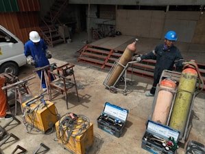 Two people wearing protective gear, including helmets, are working with welding equipment. They are surrounded by various tools and large gas cylinders on carts, likely for industrial use. The setting appears to be an industrial workshop with metal structures and a parked van in the background.
