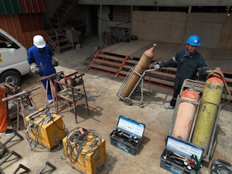 Two people wearing protective gear, including helmets, are working with welding equipment. They are surrounded by various tools and large gas cylinders on carts, likely for industrial use. The setting appears to be an industrial workshop with metal structures and a parked van in the background.