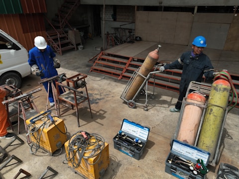 Two people wearing protective gear, including helmets, are working with welding equipment. They are surrounded by various tools and large gas cylinders on carts, likely for industrial use. The setting appears to be an industrial workshop with metal structures and a parked van in the background.