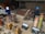 Two people wearing protective gear, including helmets, are working with welding equipment. They are surrounded by various tools and large gas cylinders on carts, likely for industrial use. The setting appears to be an industrial workshop with metal structures and a parked van in the background.