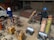 Two people wearing protective gear, including helmets, are working with welding equipment. They are surrounded by various tools and large gas cylinders on carts, likely for industrial use. The setting appears to be an industrial workshop with metal structures and a parked van in the background.