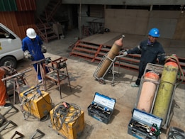Two people wearing protective gear, including helmets, are working with welding equipment. They are surrounded by various tools and large gas cylinders on carts, likely for industrial use. The setting appears to be an industrial workshop with metal structures and a parked van in the background.