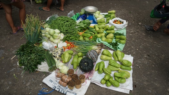An assortment of fresh vegetables displayed on the ground at a market. The collection includes green leafy vegetables, cabbage, green beans, chayote, tomatoes, and several other types of produce. Some items are placed in bags and others directly on a cloth. A weighing scale is visible among the vegetables, indicating that this is a selling point for produce.
