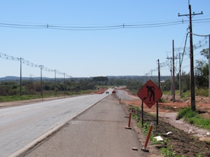 A partially constructed road with power lines running alongside it, bordered by green fields and distant trees. A warning sign indicates construction work, and a single vehicle can be seen in the distance.