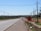 A partially constructed road with power lines running alongside it, bordered by green fields and distant trees. A warning sign indicates construction work, and a single vehicle can be seen in the distance.