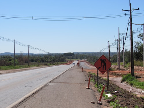 A partially constructed road with power lines running alongside it, bordered by green fields and distant trees. A warning sign indicates construction work, and a single vehicle can be seen in the distance.