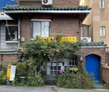 A quaint brick building with a green tiled roof, featuring a small storefront adorned with vibrant plants and flowers. A yellow sign with Korean writing is displayed above the entrance, and an air conditioning unit is visible. The entrance door is glass, revealing notices or menus. The scene is calm and inviting, with a scattering of potted plants enhancing the homey atmosphere.
