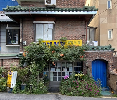 A quaint brick building with a green tiled roof, featuring a small storefront adorned with vibrant plants and flowers. A yellow sign with Korean writing is displayed above the entrance, and an air conditioning unit is visible. The entrance door is glass, revealing notices or menus. The scene is calm and inviting, with a scattering of potted plants enhancing the homey atmosphere.