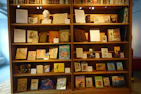 A large wooden bookshelf filled with a variety of vintage books and displayed items. The shelves are well-lit, showcasing numerous colorful book covers and some framed pages. A white sculpted bust is placed prominently among the books, adding a classical touch. Informational placards are neatly arranged alongside the books, providing context or descriptions.