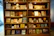 A large wooden bookshelf filled with a variety of vintage books and displayed items. The shelves are well-lit, showcasing numerous colorful book covers and some framed pages. A white sculpted bust is placed prominently among the books, adding a classical touch. Informational placards are neatly arranged alongside the books, providing context or descriptions.
