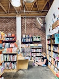 A cozy bookstore with wooden bookshelves filled with colorful books lining the walls. A woman and a child sit on the floor in a corner reading a book, accompanied by a fluffy white dog. The setting has an exposed brick wall and wooden ceiling beams, creating a warm and inviting atmosphere.