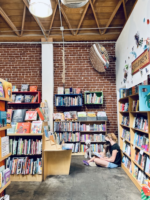 A cozy bookstore with wooden bookshelves filled with colorful books lining the walls. A woman and a child sit on the floor in a corner reading a book, accompanied by a fluffy white dog. The setting has an exposed brick wall and wooden ceiling beams, creating a warm and inviting atmosphere.