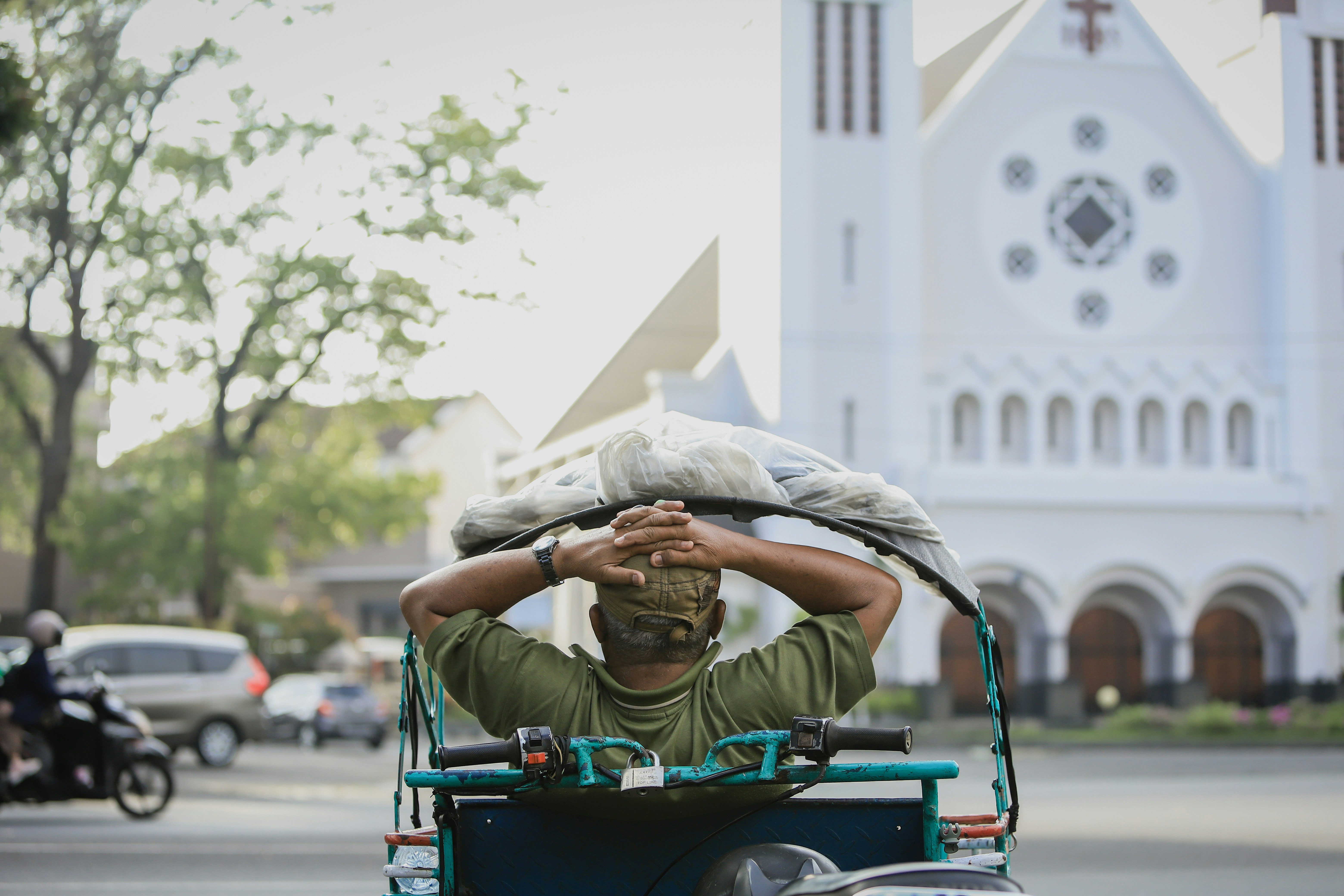 a person riding a scooter with an umbrella on the head, Rickshaw rider sitting holding his head with two hands view from behind and church view