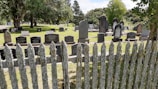 A cemetery with numerous headstones arranged in rows on well-maintained grass. A weathered wooden fence covered in moss or lichen stands in the foreground. Mature trees provide shade and a serene atmosphere.