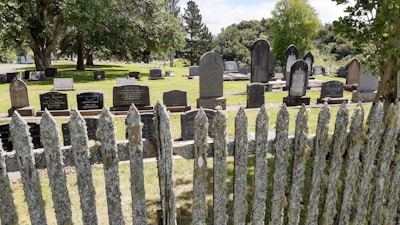A cemetery with numerous headstones arranged in rows on well-maintained grass. A weathered wooden fence covered in moss or lichen stands in the foreground. Mature trees provide shade and a serene atmosphere.