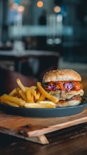 A close-up of a juicy burger with colorful toppings, including lettuce and purple cabbage, next to a serving of golden french fries. The dish is on a dark plate placed on a wooden board, with a blurred restaurant setting in the background.