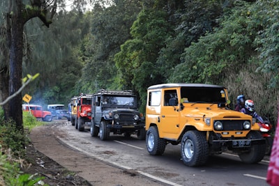 A line of off-road vehicles, including a prominent yellow Jeep, is parked on a narrow road surrounded by lush greenery. Several motorcycles and riders wearing helmets are also visible. The scene suggests a group adventure in a rural or forested area.