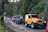 A line of off-road vehicles, including a prominent yellow Jeep, is parked on a narrow road surrounded by lush greenery. Several motorcycles and riders wearing helmets are also visible. The scene suggests a group adventure in a rural or forested area.