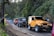 A line of off-road vehicles, including a prominent yellow Jeep, is parked on a narrow road surrounded by lush greenery. Several motorcycles and riders wearing helmets are also visible. The scene suggests a group adventure in a rural or forested area.