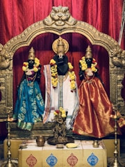 Three intricately crafted black statues adorned with colorful clothing and garlands stand within a beautifully ornate golden frame. The statues are decorated with bright floral garlands and appear to be part of a religious or cultural setting. The background features rich red drapery that enhances the vibrancy of the attire and decorations.