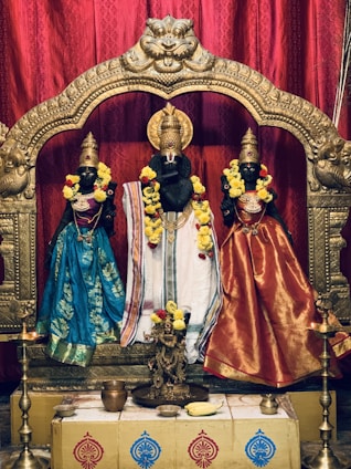 Three intricately crafted black statues adorned with colorful clothing and garlands stand within a beautifully ornate golden frame. The statues are decorated with bright floral garlands and appear to be part of a religious or cultural setting. The background features rich red drapery that enhances the vibrancy of the attire and decorations.