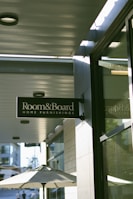 A street view displaying a sign for 'Room & Board Home Furnishings' hanging above a sidewalk. The sign is mounted on the side of a building with a reflective glass window. Below, a patio umbrella is partially visible, suggesting an outdoor seating area. In the background, other buildings and a lamppost are faintly discernible, indicating an urban environment. Soft sunlight casts shadows, creating a serene atmosphere.