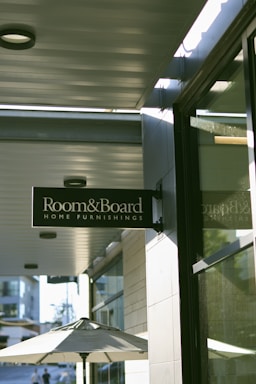 A street view displaying a sign for 'Room & Board Home Furnishings' hanging above a sidewalk. The sign is mounted on the side of a building with a reflective glass window. Below, a patio umbrella is partially visible, suggesting an outdoor seating area. In the background, other buildings and a lamppost are faintly discernible, indicating an urban environment. Soft sunlight casts shadows, creating a serene atmosphere.