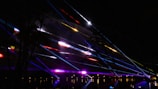 Colorful laser beams are projected across a dark sky, creating vibrant streaks of light. The beams are in various colors including red, blue, yellow, and purple, intersecting one another in a dynamic display. Silhouettes of trees and some faint outlines of people are visible, adding depth to the scene.