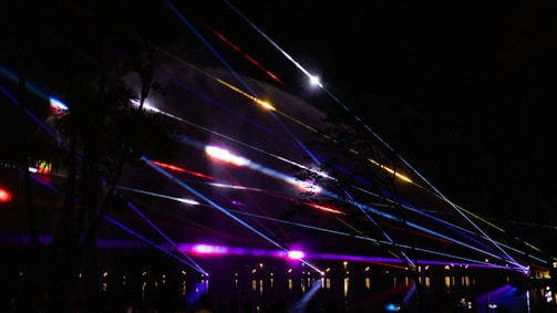 Colorful laser beams are projected across a dark sky, creating vibrant streaks of light. The beams are in various colors including red, blue, yellow, and purple, intersecting one another in a dynamic display. Silhouettes of trees and some faint outlines of people are visible, adding depth to the scene.