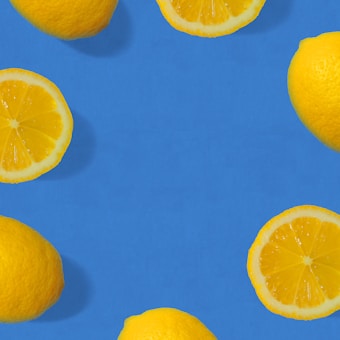 Whole and sliced lemons are arranged on a bright blue background, forming a pattern with a circular empty space in the center. The vibrant contrast between the yellow lemons and the blue backdrop creates a visually striking composition.