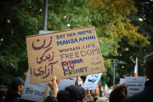 A large protest with people holding signs displaying messages in multiple languages, focusing on a specific person's name and themes of freedom and rights. The protest appears to be outdoors with trees in the background.