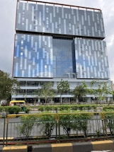 A modern high-rise building with a glass facade reflecting the sky. The building has a geometric pattern and is surrounded by greenery and a road with vehicles, including an auto-rickshaw.