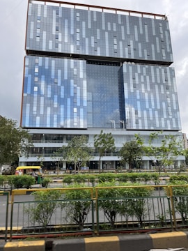 A modern high-rise building with a glass facade reflecting the sky. The building has a geometric pattern and is surrounded by greenery and a road with vehicles, including an auto-rickshaw.