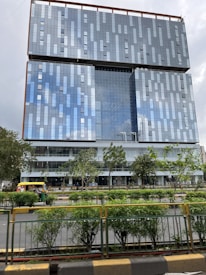 A modern high-rise building with a glass facade reflecting the sky. The building has a geometric pattern and is surrounded by greenery and a road with vehicles, including an auto-rickshaw.