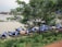 A group of traditional wooden boats with blue tarpaulins bearing a Pepsi logo are docked along a riverbank. The boats are positioned in a row next to a lush green area with trees and grass. The river appears calm, and there is a bridge visible in the background.