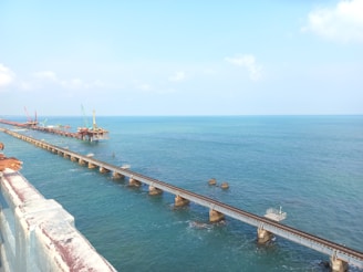 A long railway bridge extends over a calm sea with clear blue water, supported by numerous concrete pillars. Construction cranes and machinery are visible in the distance on a separate section of the bridge.