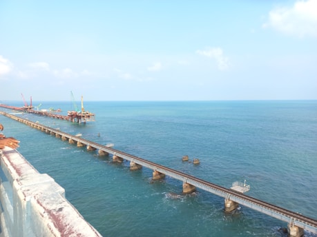 A long railway bridge extends over a calm sea with clear blue water, supported by numerous concrete pillars. Construction cranes and machinery are visible in the distance on a separate section of the bridge.