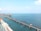 A long railway bridge extends over a calm sea with clear blue water, supported by numerous concrete pillars. Construction cranes and machinery are visible in the distance on a separate section of the bridge.