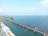 A long railway bridge extends over a calm sea with clear blue water, supported by numerous concrete pillars. Construction cranes and machinery are visible in the distance on a separate section of the bridge.