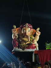 A large statue of Ganesha, intricately decorated, is suspended by a crane in a nighttime setting. The crowd below captures the moment using mobile phones. The statue is adorned with floral garlands and vibrant clothing, and bright spotlights create dramatic illumination.