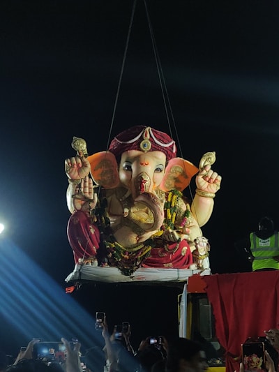 A large statue of Ganesha, intricately decorated, is suspended by a crane in a nighttime setting. The crowd below captures the moment using mobile phones. The statue is adorned with floral garlands and vibrant clothing, and bright spotlights create dramatic illumination.