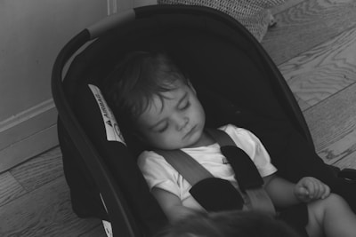 A young child is peacefully asleep in a car seat. The child is wearing a white shirt and is buckled in the seat, which is placed on a wooden floor. Light gently illuminates the scene, highlighting the child's relaxed expression.